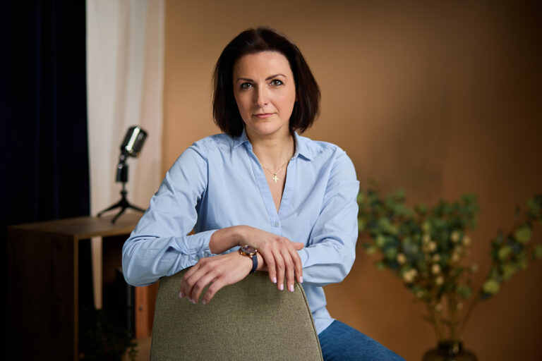 Femeie în cămașă albastru deschis, relaxată pe scaun, într-un decor cald de studio